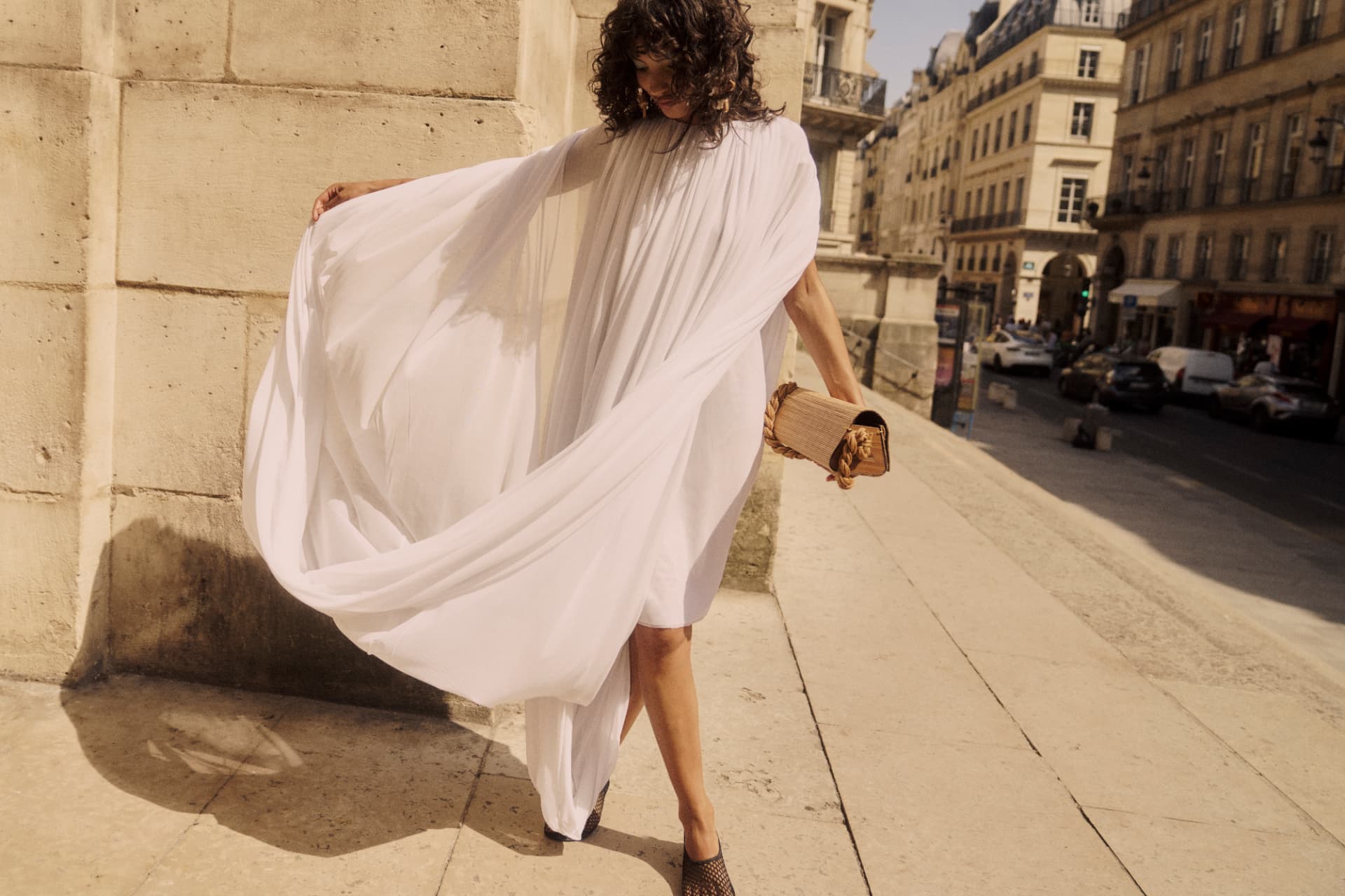 Model in a flowing white pleated dress with cape sleeves walking a sunlit Parisian street, carrying a woven clutch